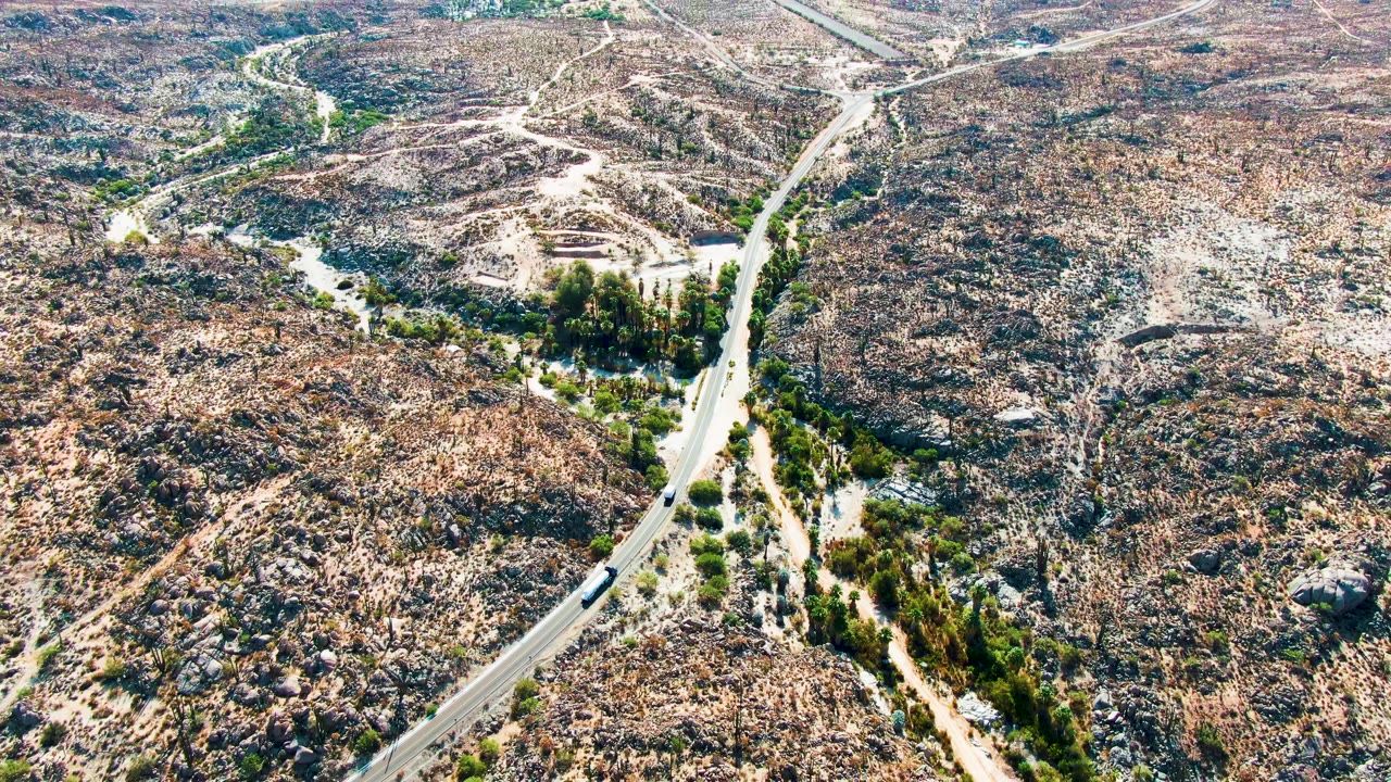 Aerial view in Catavina BC, Mexico with the vehicle on the lower road segment, multiple pale dirt paths across the rocky slope, scattered vegetation, and no visible people.