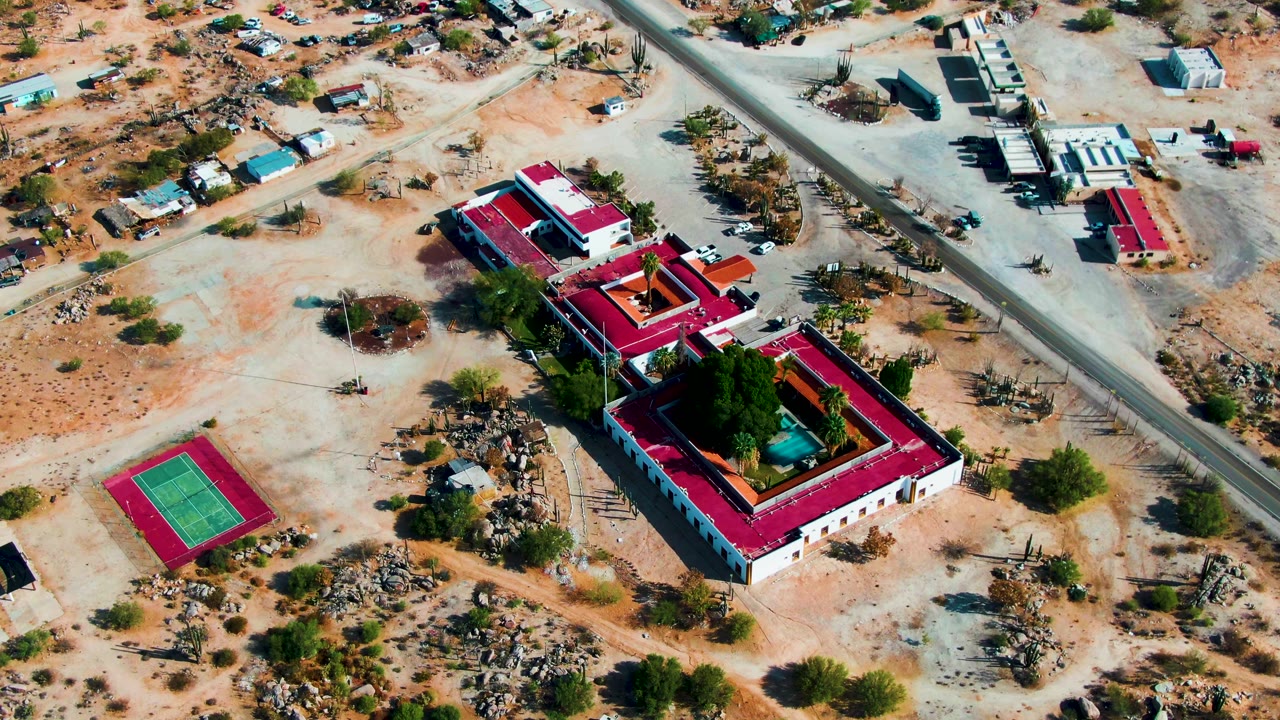 Aerial frame over Catavina BC, Mexico showing a red-roofed compound, green tennis court, sandy lots, sparse shrubs, nearby roads, and no visible people.