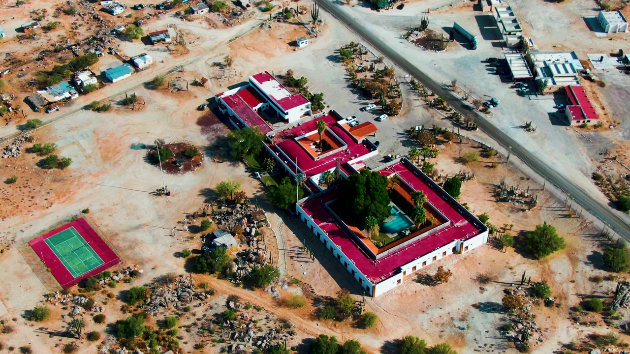 Second aerial frame in Catavina BC, Mexico with the red-roofed buildings larger in view, the tennis court at left, roadways above, parked vehicles, and no visible people.