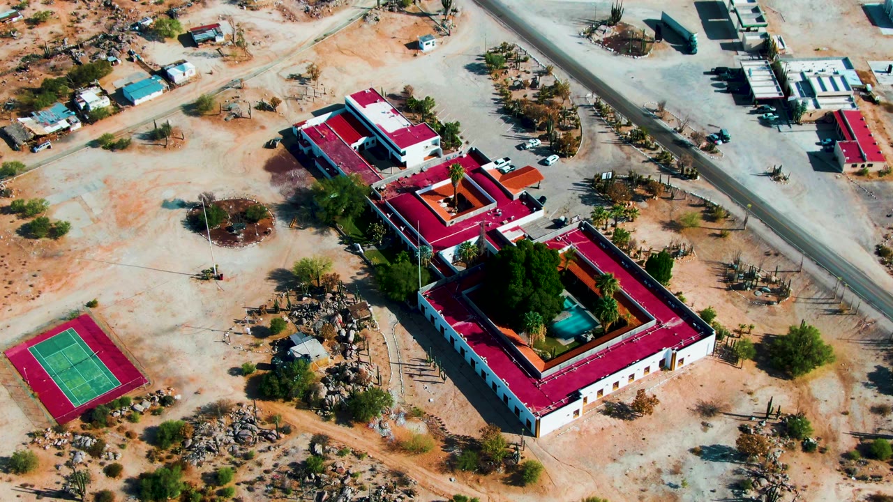 Aerial view over Catavina BC, Mexico showing the walled red-roofed compound, interior trees and blue pool-like feature, a tennis court, surrounding sand, roads, and no visible people.