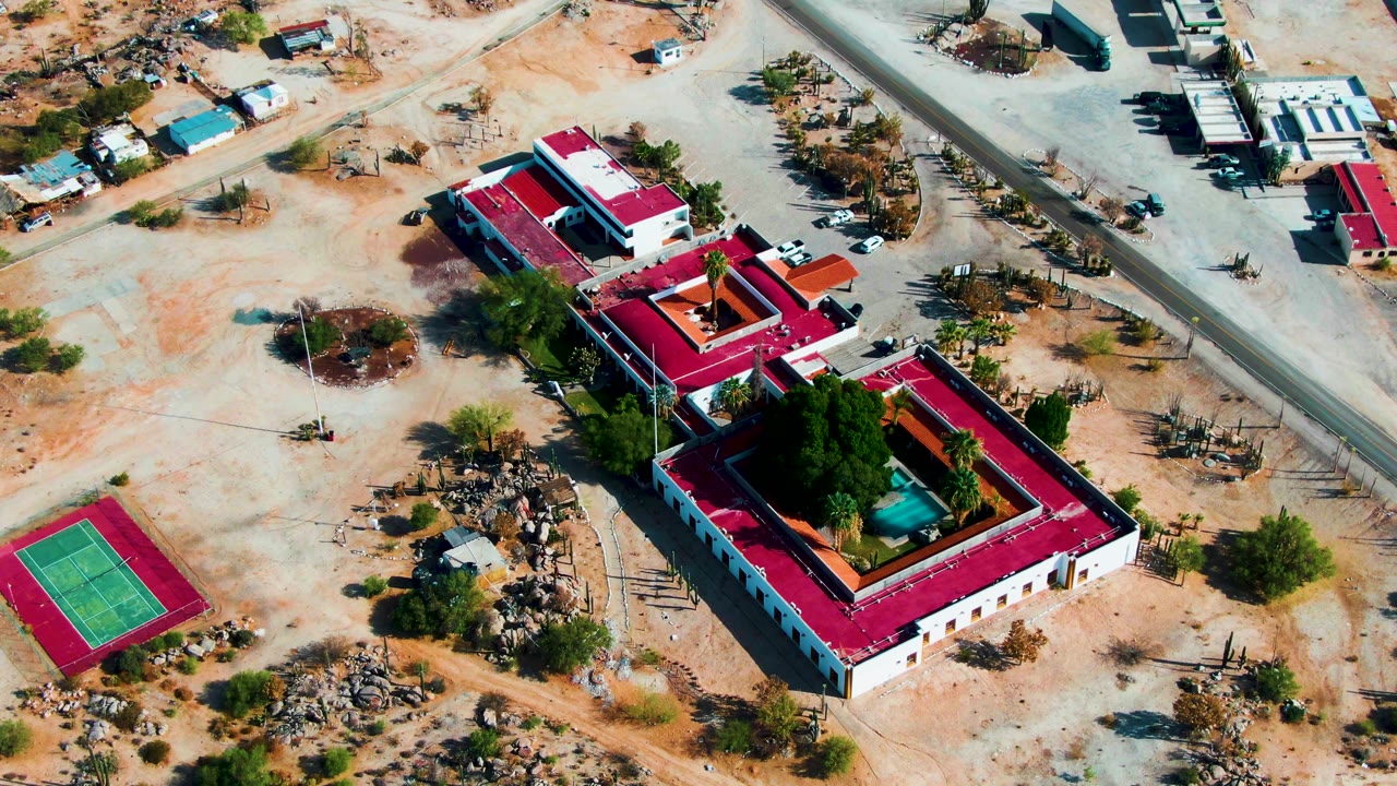 Aerial pass over red-roofed buildings, a tennis court, sandy lots, and roadside structures.