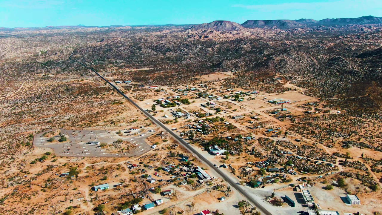 Early aerial frame over Catavina BC Mexico showing a diagonal paved road, sandy settlement, airstrip area, sparse vegetation, distant mountains, clear blue sky, and no visible people.