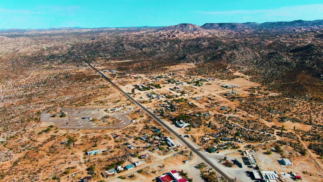 Aerial frame over Catavina BC Mexico with the paved road leading from the lower frame toward the horizon, low buildings and vehicles on sandy lots, an airstrip, rocky hills, and no visible people.