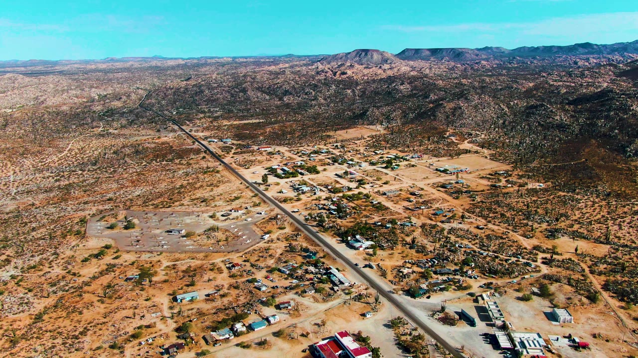 Mid-clip aerial frame in Catavina BC Mexico showing the airstrip and settlement from above, with small structures, vehicles, scrub vegetation, brown desert ground, mountains, and no visible people.