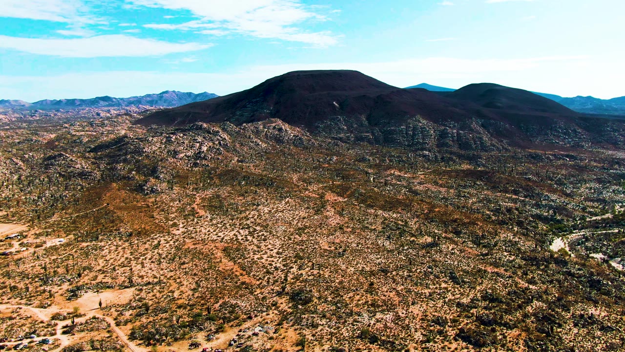 Opening aerial view in Catavina BC Mexico showing a pale dirt road with a small vehicle crossing scrubby desert terrain, dark rounded mountains in the background, and no visible people.