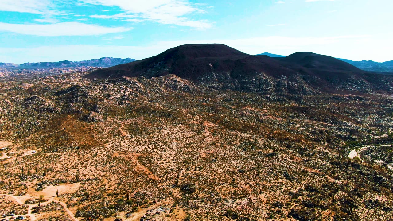 Wide aerial frame over Catavina BC Mexico desert, with the dirt road and tiny vehicle set low in the scrubland while the dark mountain ridge spans the horizon, and no visible people.