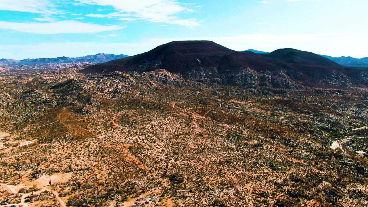 Aerial view in Catavina BC Mexico with the road threading through sparse desert vegetation, a small vehicle on the lower left road area, rounded dark mountains behind, and no visible people.