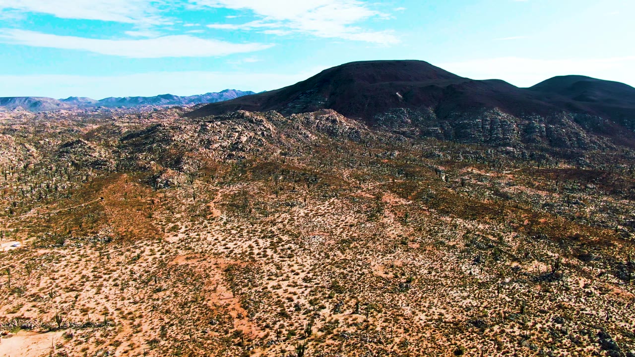 Opening frame over Catavina BC Mexico shows scrub desert and pale dirt tracks leading toward rocky hills, dark rounded mountains, and bright sky with no visible people.