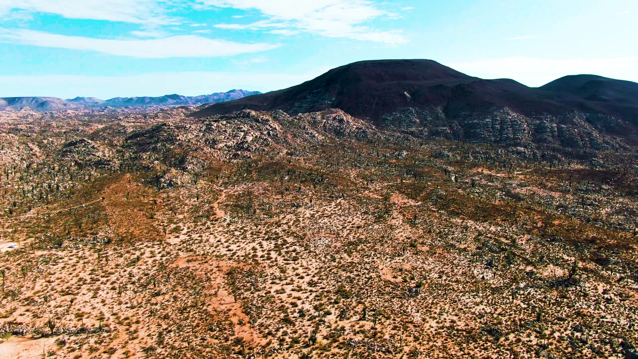 At 0:01, the Catavina BC Mexico aerial view holds on sparse desert vegetation, rough tan ground, rocky midground ridges, and dark mountains with no visible people.