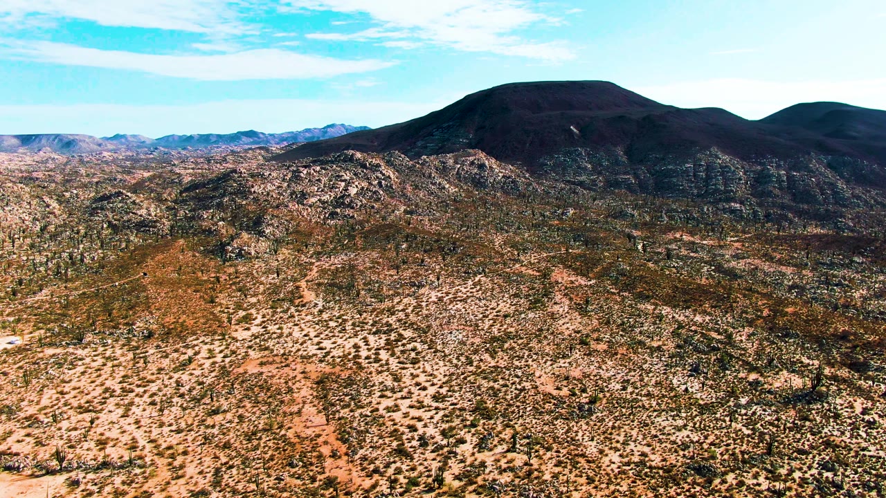 At 0:02, the frame shows a faint dirt road through the Catavina BC Mexico desert basin, with scrub, rocky slopes, and a dark mountain ridge under clear daylight with no visible people.