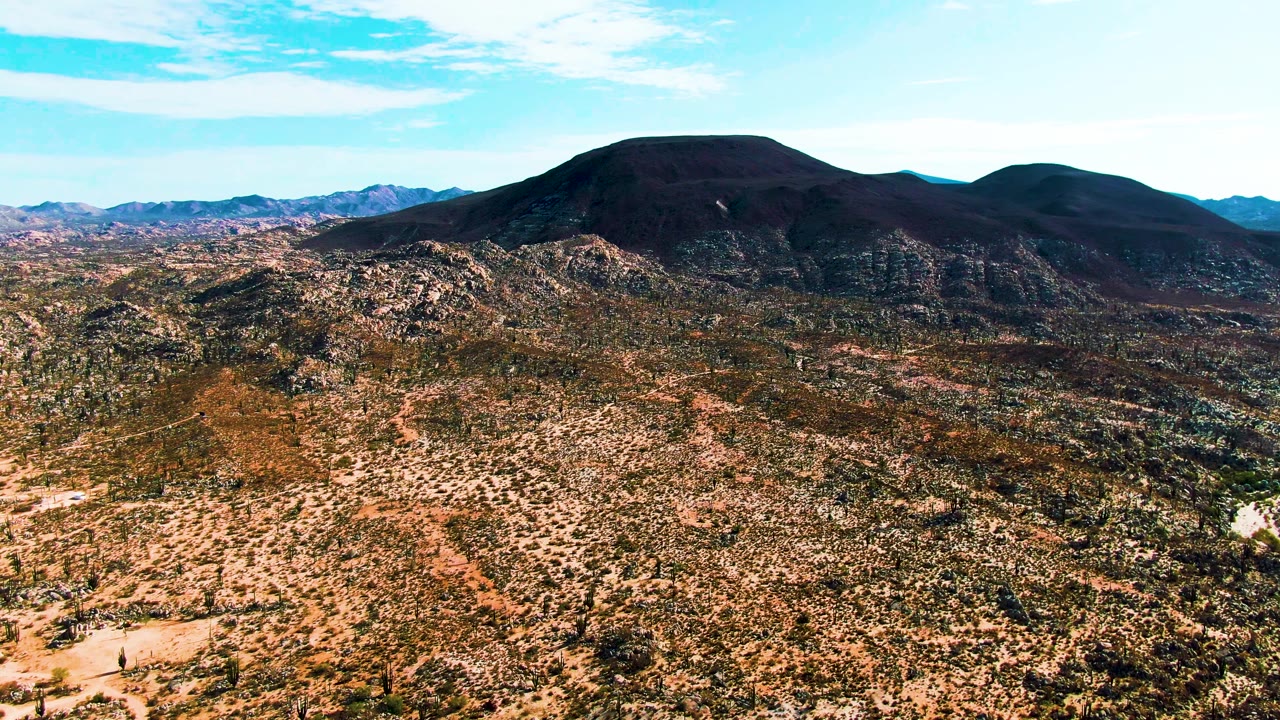 Aerial view over Catavina BC Mexico desert with rocky tan terrain in the foreground, dark rounded mountains on the horizon, thin clouds in a blue sky, and no visible people.