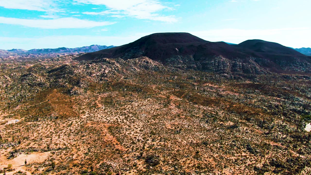 Catavina BC Mexico aerial frame with mottled desert scrub and pale rock patches stretching toward a broad dark mountain ridge beneath a bright blue sky, with no visible people.