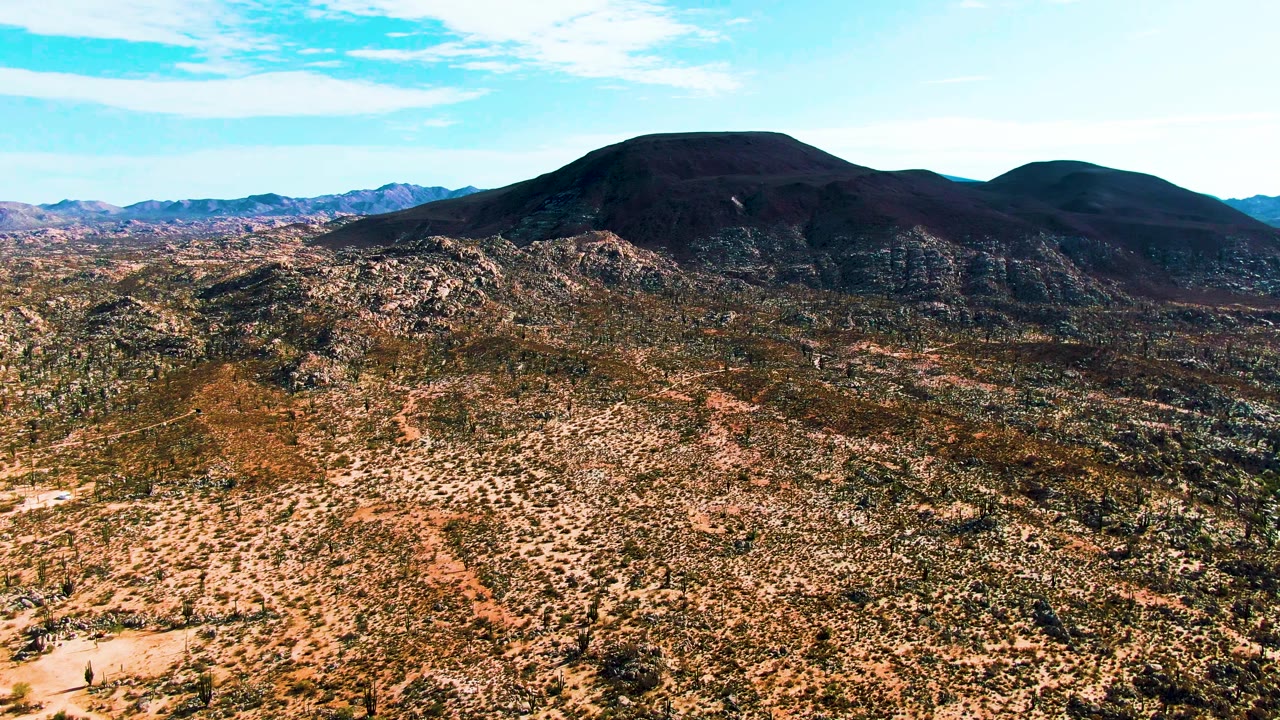 Aerial view in Catavina BC Mexico showing dense speckled scrub across dry rocky ground, low hills in the midground, distant mountains, pale cloud streaks, and no visible people.