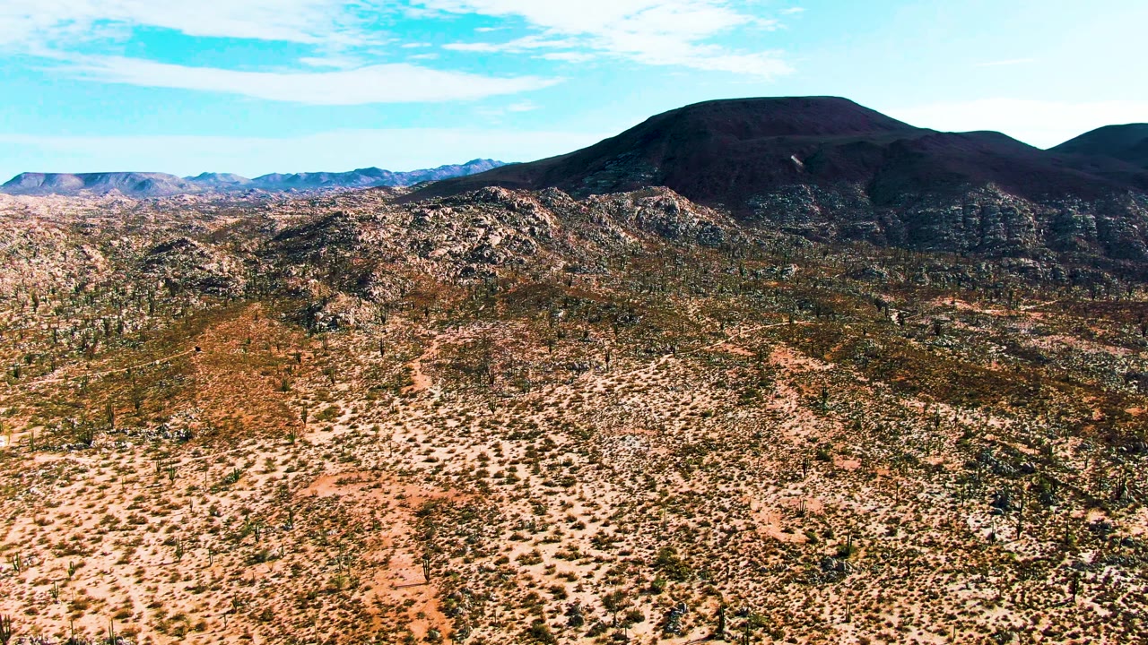 Wide aerial view over Catavina BC Mexico desert with scattered shrubs, rocky ridges, a dark mountain on the horizon, and no visible people.