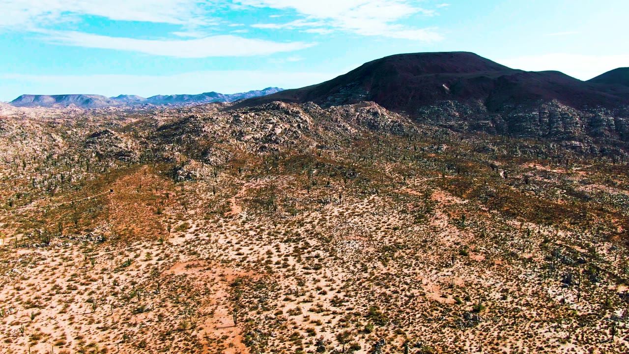 Aerial frame from Catavina BC Mexico showing brown scrubland in the foreground, pale rocky hills in the middle distance, a dark mountain ridge, and no visible people.