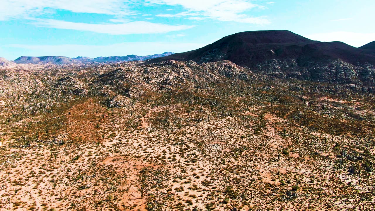 Catavina BC Mexico aerial view with textured desert shrubs, exposed rocks across the ridgeline, a broad dark mountain behind them, and no visible people.