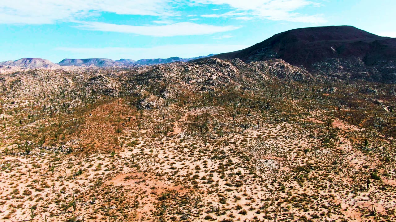 Early frame of Catavina BC Mexico desert with sandy foreground scrub, scattered cacti, rocky midground hills, distant mountains, and no visible people.