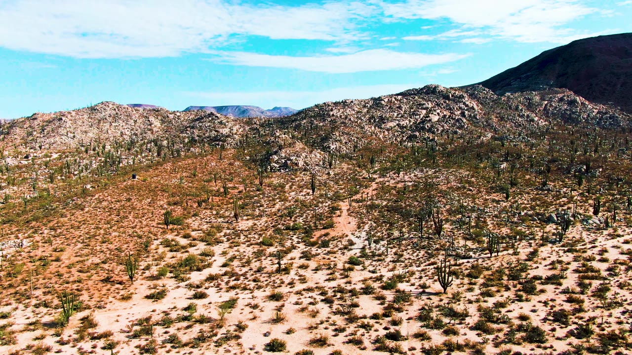 Opening frame of Catavina, Mexico desert terrain with scattered cacti across sandy scrub, rocky hills in back, a dark mountain slope at upper right, and no visible people.