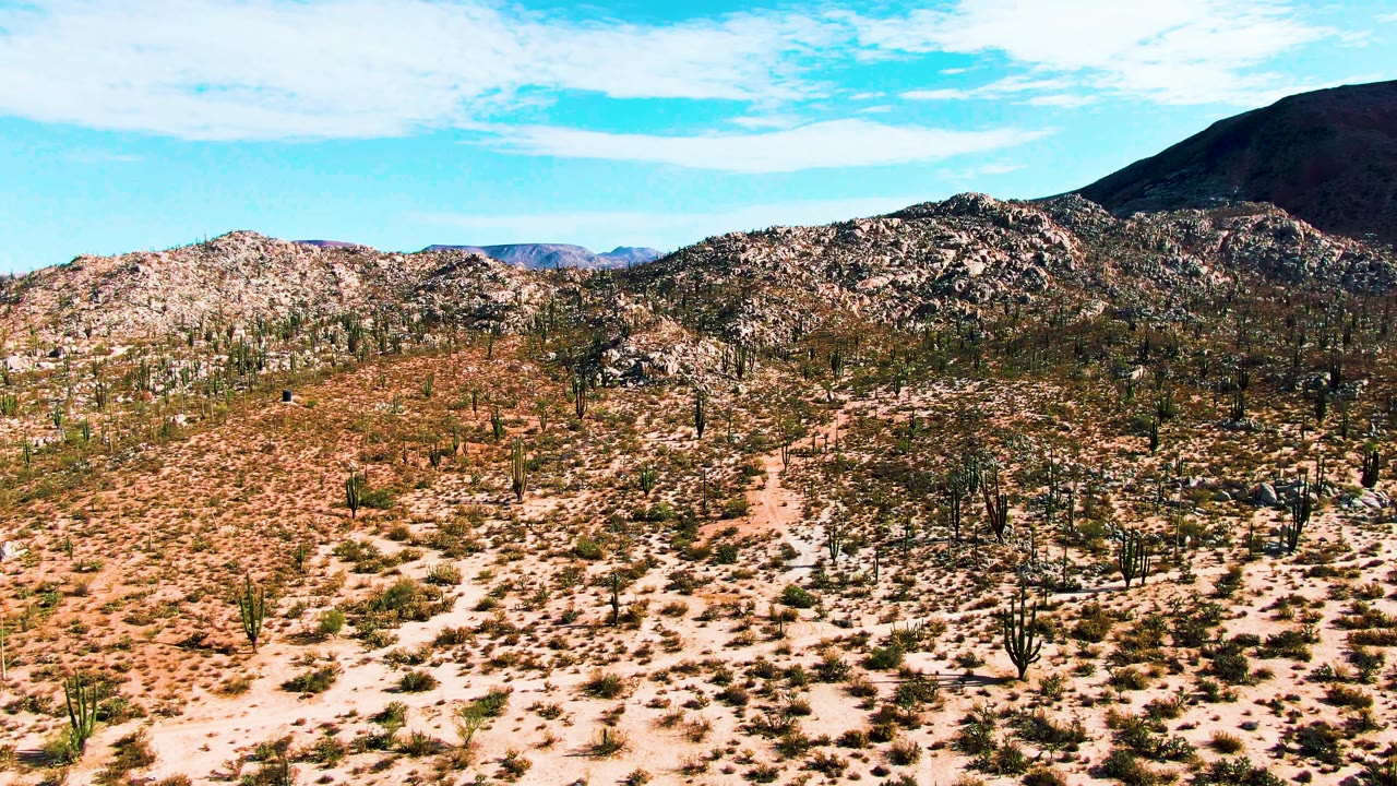 Catavina, Mexico desert scene one second in, showing a central rocky rise, saguaro-like cacti and shrubs over pale sand, distant hills, blue sky, and no visible people.