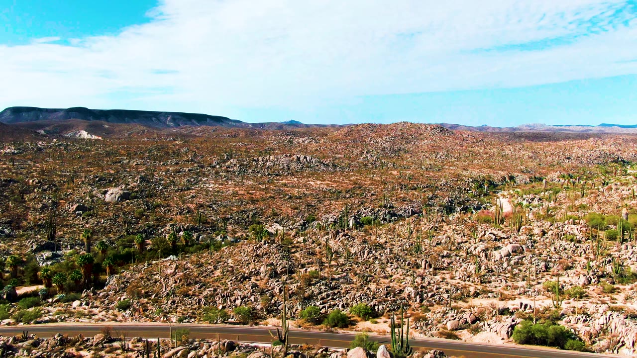 Later frame of Catavina BC Mexico with the paved road along the bottom, rocky desert and tall cacti stretching to distant hills, and no visible people.