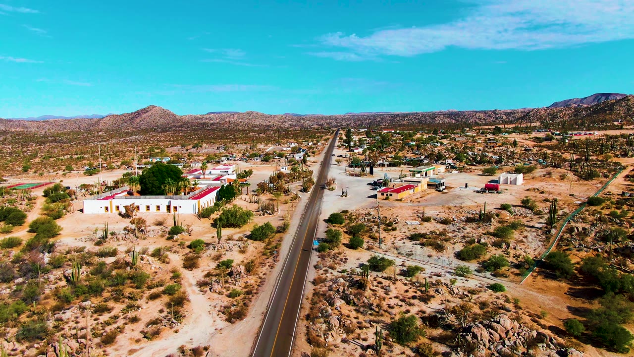 At 0:00, an aerial view in Catavina BC Mexico looks along a paved desert road with low red-roofed buildings, sandy scrub, scattered cacti, distant hills, and no visible people.