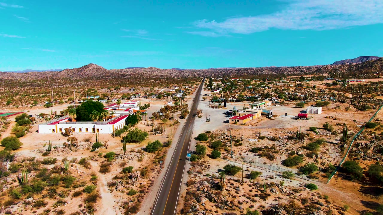 At 0:01, the Catavina BC Mexico drone view centers the straight road as roadside buildings, dry vegetation, cacti, rocky lots, and low mountains fill the frame with no visible people.
