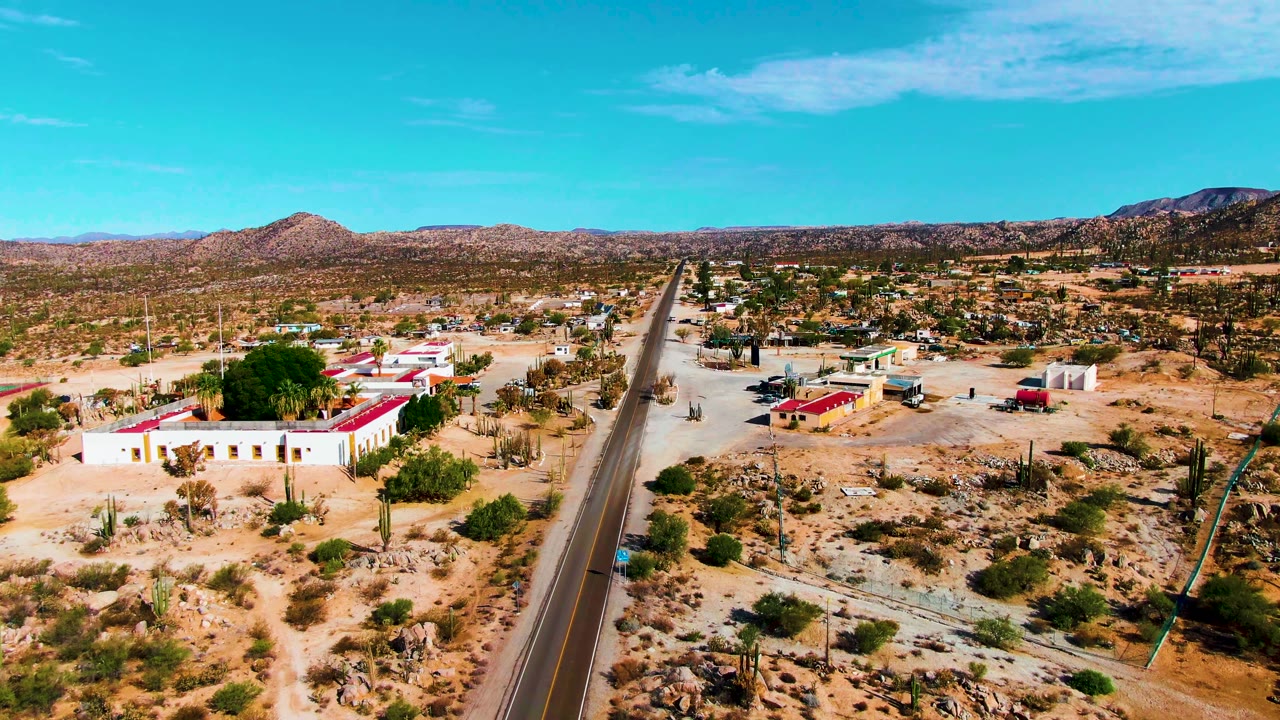 At 0:02, the aerial view shows the road passing between a white building with a red roof, small outbuildings, cactus-dotted sandy terrain, distant hills, and no visible people.