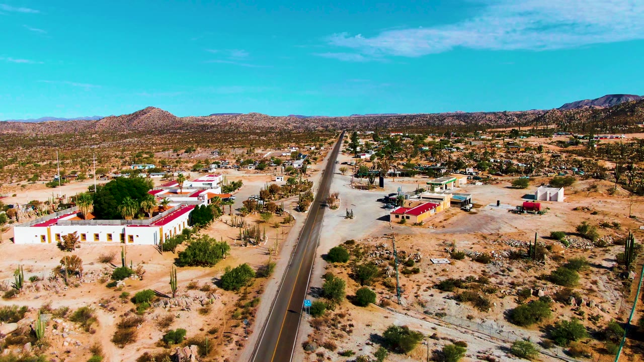 Wide aerial view of a desert road with cacti and red-roofed buildings.
