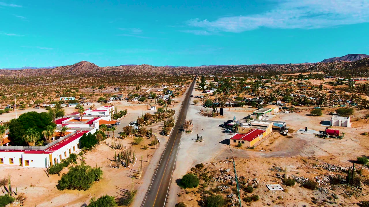 Opening aerial frame over Catavina BC Mexico with a straight road, red-roofed roadside buildings, sandy lots, cacti, distant hills, and no visible people.