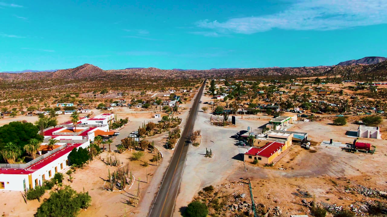 Catavina BC Mexico aerial view with the paved road running through the middle, red-roofed buildings to the left, scattered vehicles and cacti nearby, and no visible people.