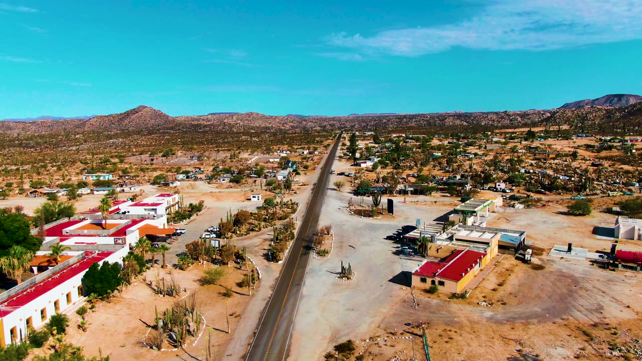 Catavina BC Mexico frame showing a straight road, red-roofed buildings in the lower left and right, scattered cacti, pale sand, low mountains, and no visible people.