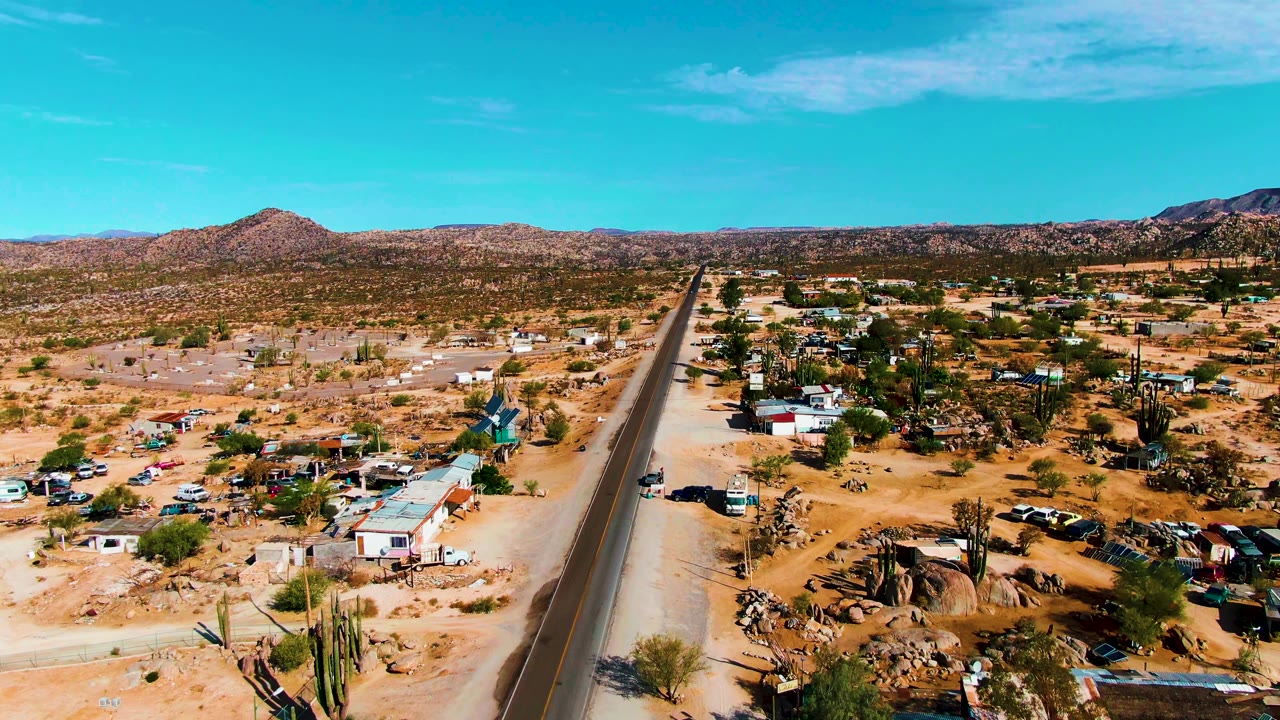 Opening aerial frame over Catavina BC Mexico shows a paved road beside scattered desert homes, tall cacti, parked vehicles, rocky hills, blue sky, and no visible people.