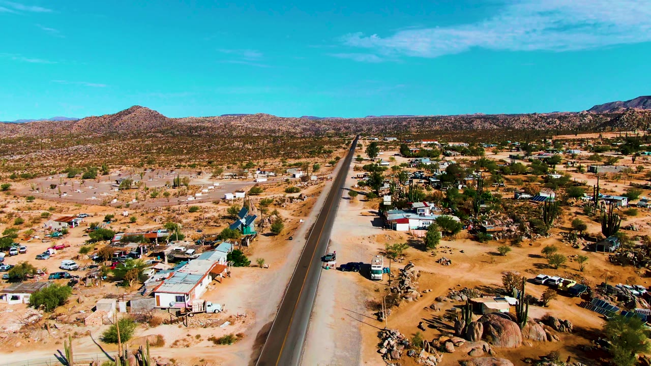 Drone view along a paved road through desert homes and cactus-covered ground.