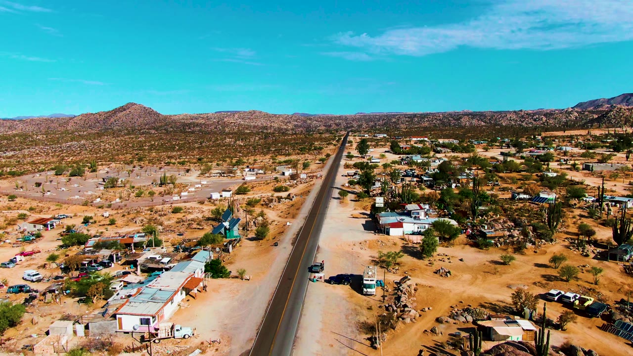 Catavina BC Mexico aerial frame shows the road cutting through dry terrain, with homes and vehicles to either side, cacti near the roadside, mountains beyond, and no visible people.
