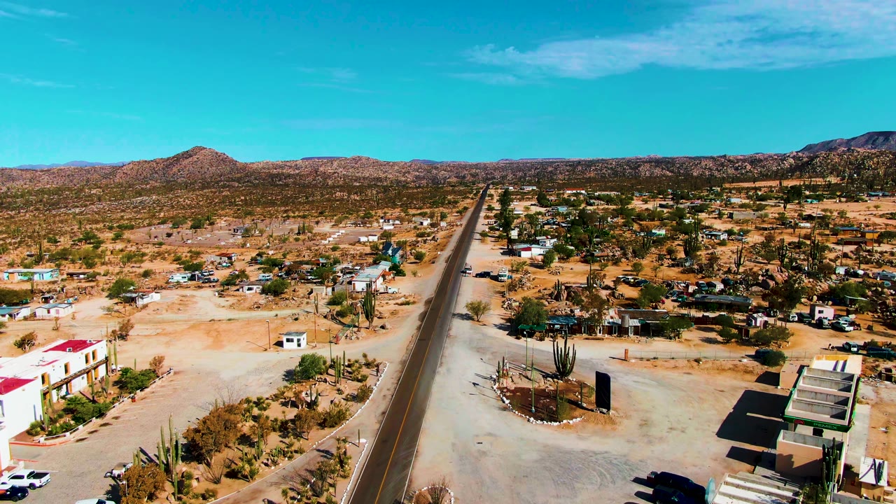 Aerial view over Catavina BC Mexico showing a paved road leading into sandy desert, scattered homes, tall cacti, distant mountains, and no visible people.