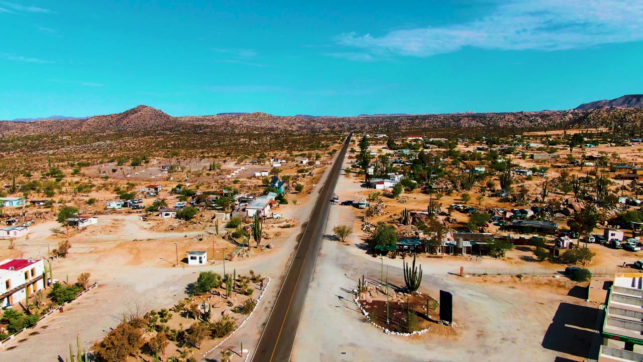 High aerial frame in Catavina BC Mexico with the paved road near center, roadside buildings, cactus clusters, sandy lots, distant hills, and no visible people.