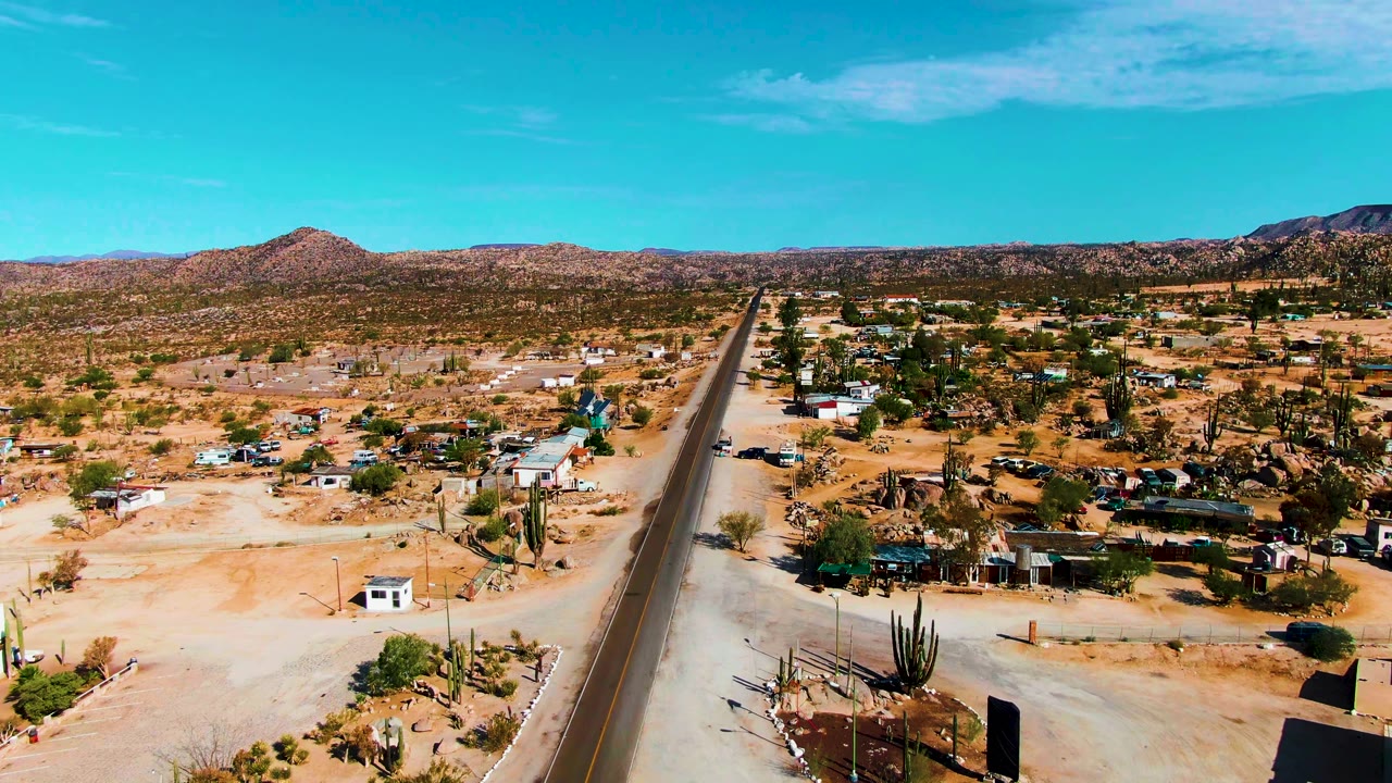 Aerial view over Catavina BC Mexico with a straight road crossing the frame, small homes and vehicles on sandy ground, cactus stands, mountains, and no visible people.