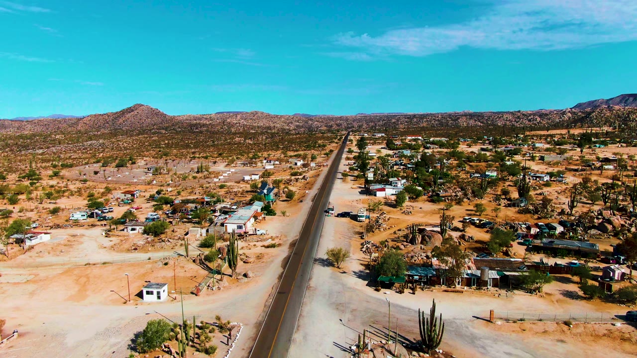 Aerial desert settlement with road, homes, cacti, and open sandy ground.