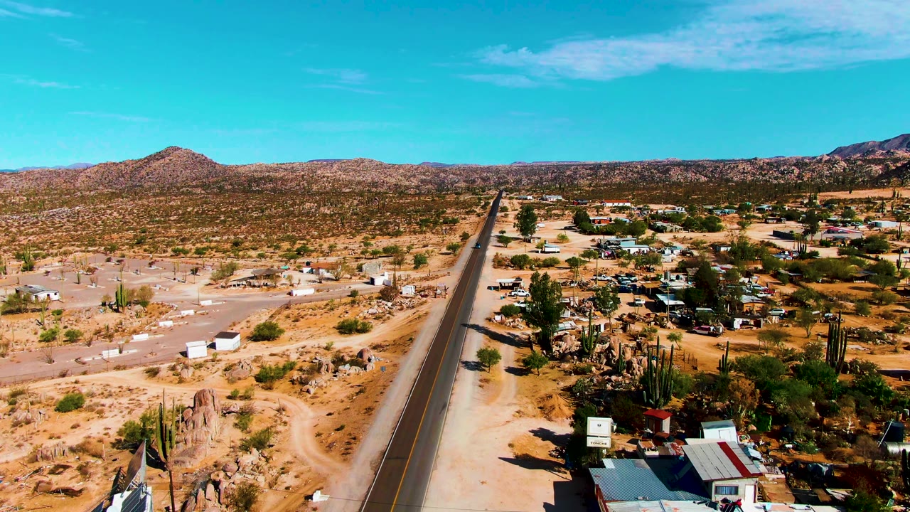 Aerial view in Catavina BC, Mexico showing a straight desert road, sandy lots, small dwellings, cacti, rocky ground, distant mountains, blue sky, and no visible people.