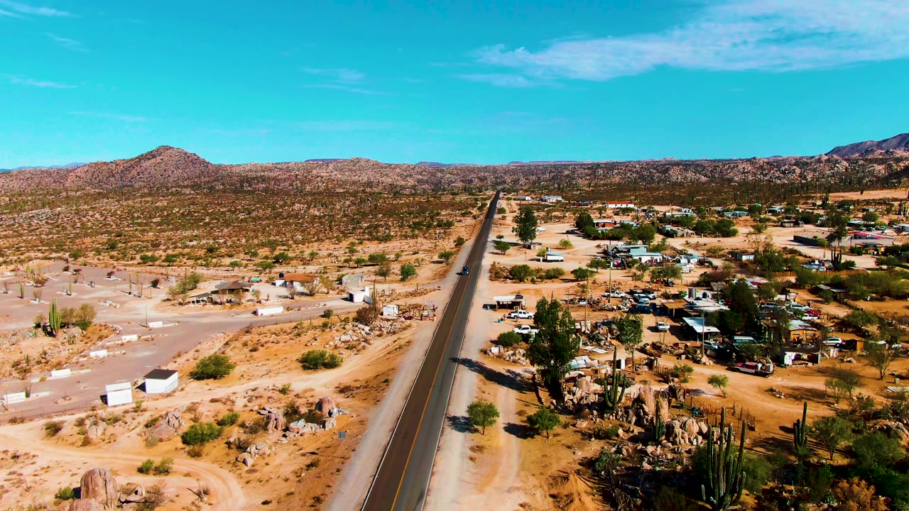 High aerial view in Catavina BC, Mexico showing the paved road running through sandy desert, a cluster of dwellings to one side, tall cacti, distant ridges, and no visible people.