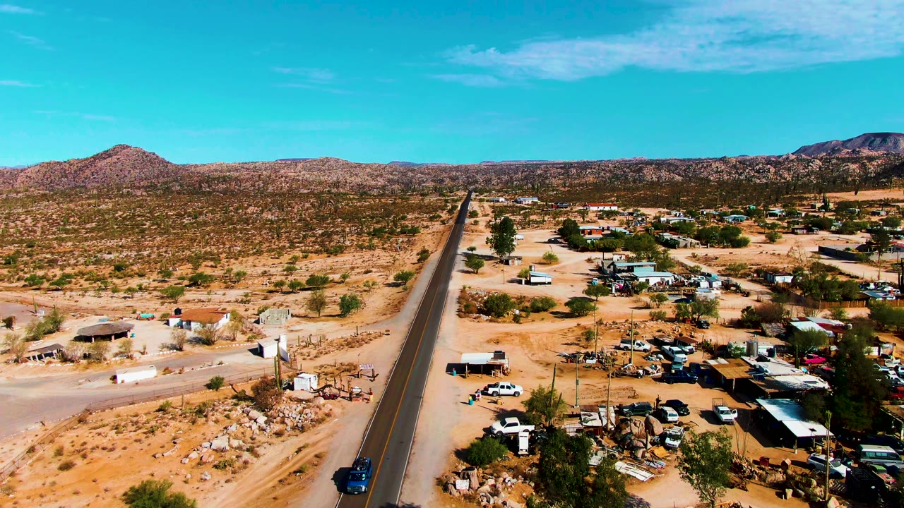Aerial frame from Catavina BC Mexico showing a straight paved road, sandy lots, low roadside buildings, parked vehicles, scrub vegetation, distant hills, blue sky, and no visible people.