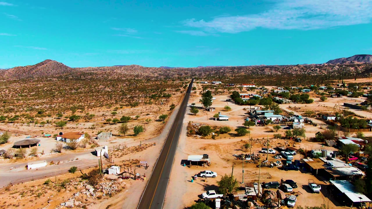 Catavina BC Mexico aerial view with the road centered toward the horizon, small structures and parked vehicles to the right, open scrub desert to the left, rocky hills in the distance, and no visible people.