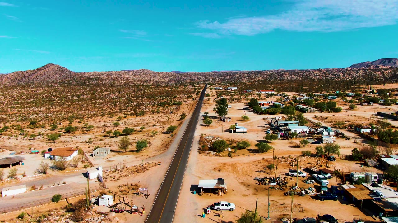 Forward aerial view over Catavina BC Mexico with a dark paved road, clustered small buildings, light-colored vehicles, dry ground, sparse green shrubs, distant mountain ridges, and no visible people.