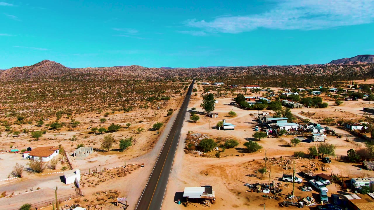 Road through desert settlement with parked vehicles and low buildings under blue sky.