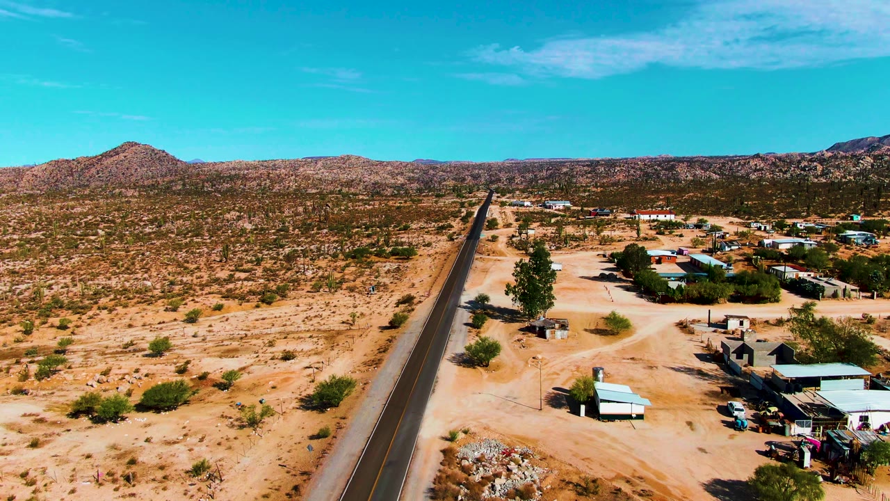 Aerial frame over Catavina BC, Mexico showing a paved road cutting through sandy desert with low buildings to the right, rocky hills in the distance, and no visible people.