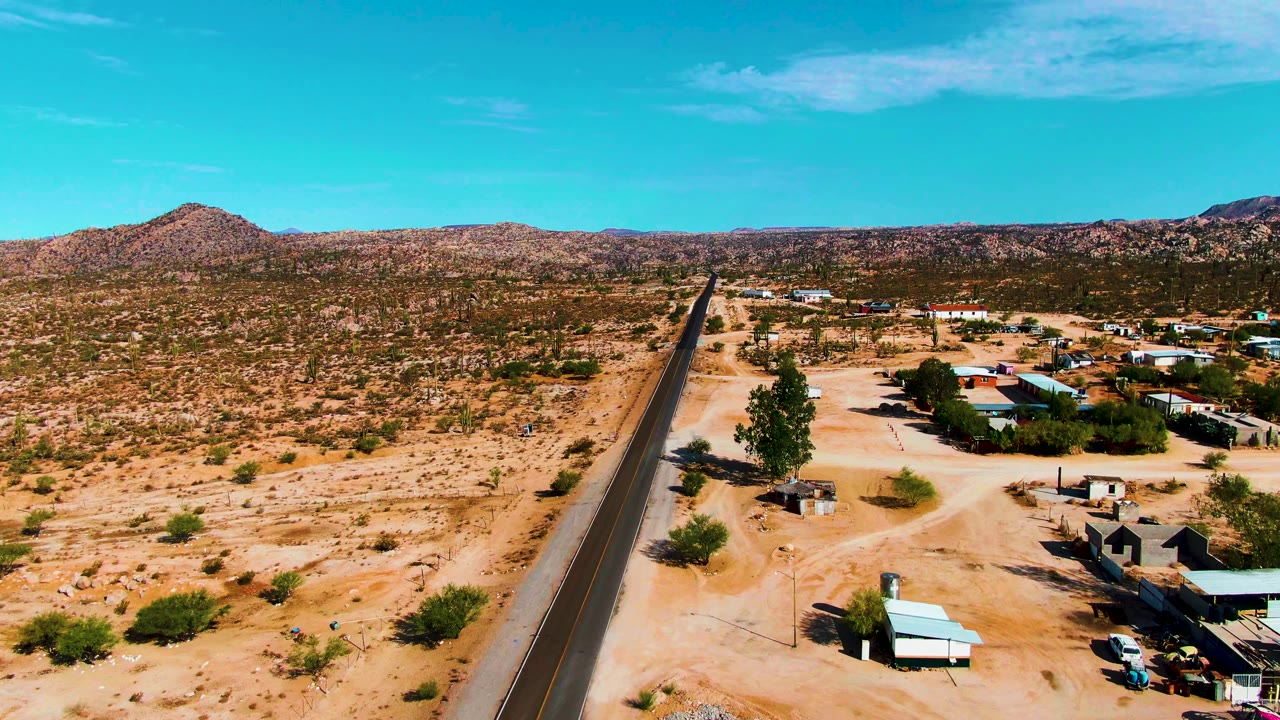 Aerial view in Catavina BC, Mexico with the highway slightly left of center, small structures and open yards to the right, sandy scrubland around it, and no visible people.