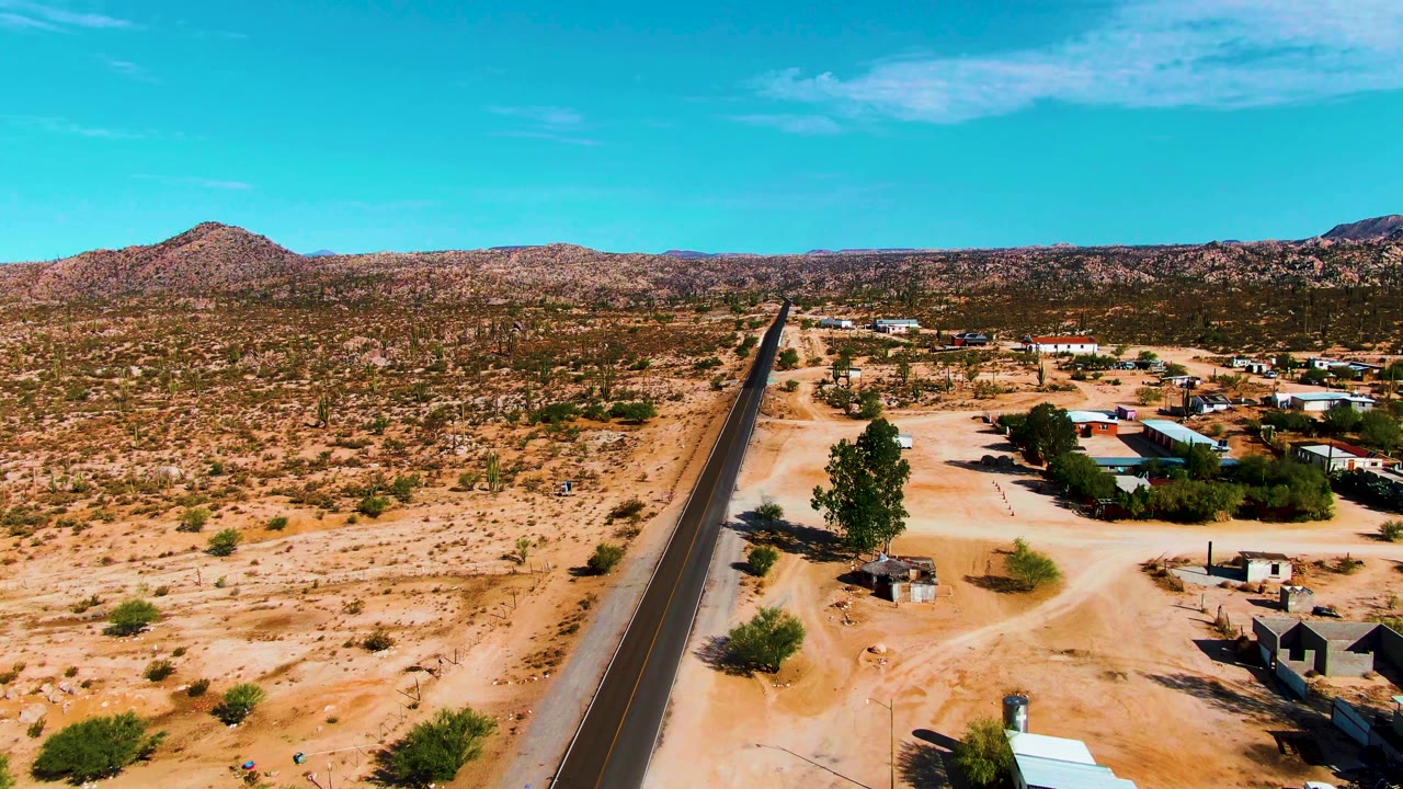 Catavina BC, Mexico aerial frame showing the road running toward the horizon, a cluster of low buildings on the right, dirt access tracks, scrub vegetation, and no visible people.