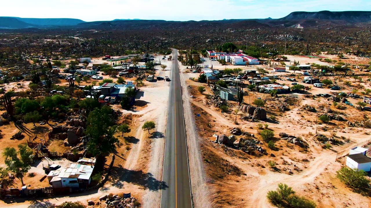 Aerial frame over Catavina, Mexico showing a paved road centered between sandy lots, scattered buildings, cacti, parked vehicles, and distant hills, with no visible people.
