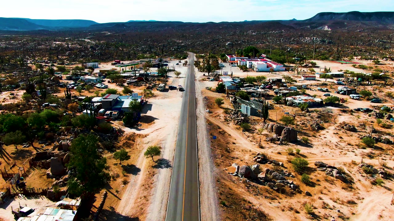 A high view in Catavina, Mexico shows the paved road running upward through sandy ground with buildings and parked vehicles on both sides, plus cacti and low hills beyond, with no visible people.