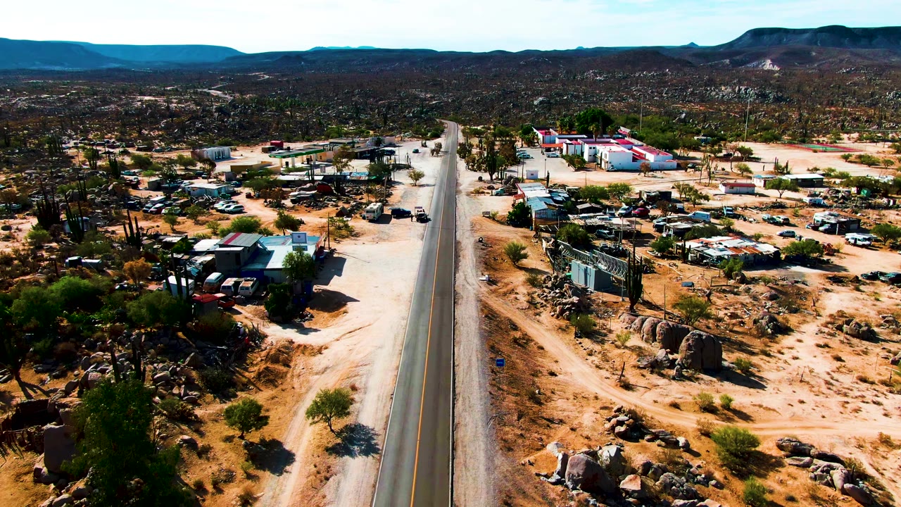 The Catavina, Mexico aerial view shows the road bordered by sandy tracks, small structures, clustered vehicles, tall cacti, boulder patches, and a broad desert horizon, with no visible people.